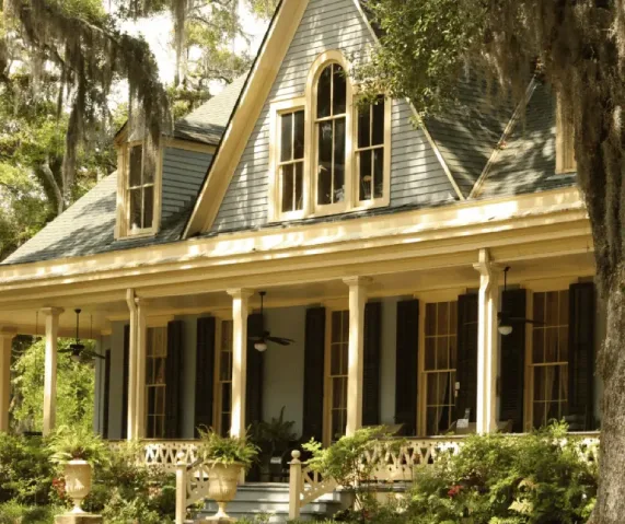 Charming Southern-style home with tall windows, wooden shutters, and a spacious wraparound porch surrounded by lush greenery.