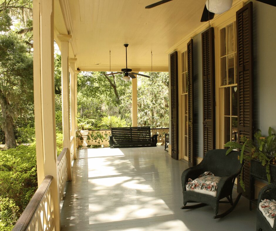 Classic wraparound porch with ceiling fans, rocking chairs, and a hanging porch swing, framed by trees and greenery.